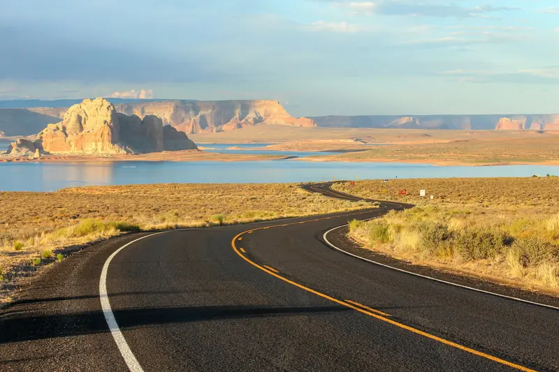 A two-lane desert highway curving past red rock buttes and a lake.
